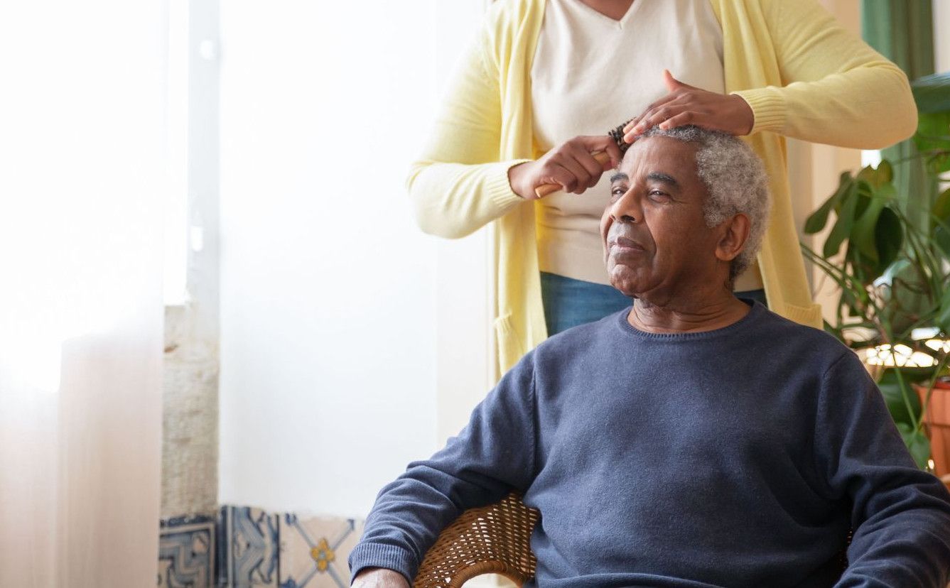 A caregiver combing an elderly man's hair near a sunlit window indoors.