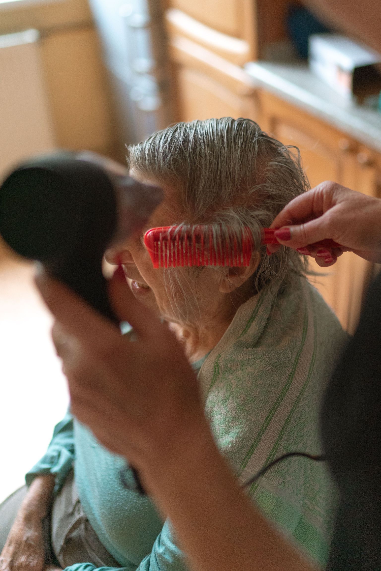 A caregiver assists a senior adult with hair drying and combing at home.
